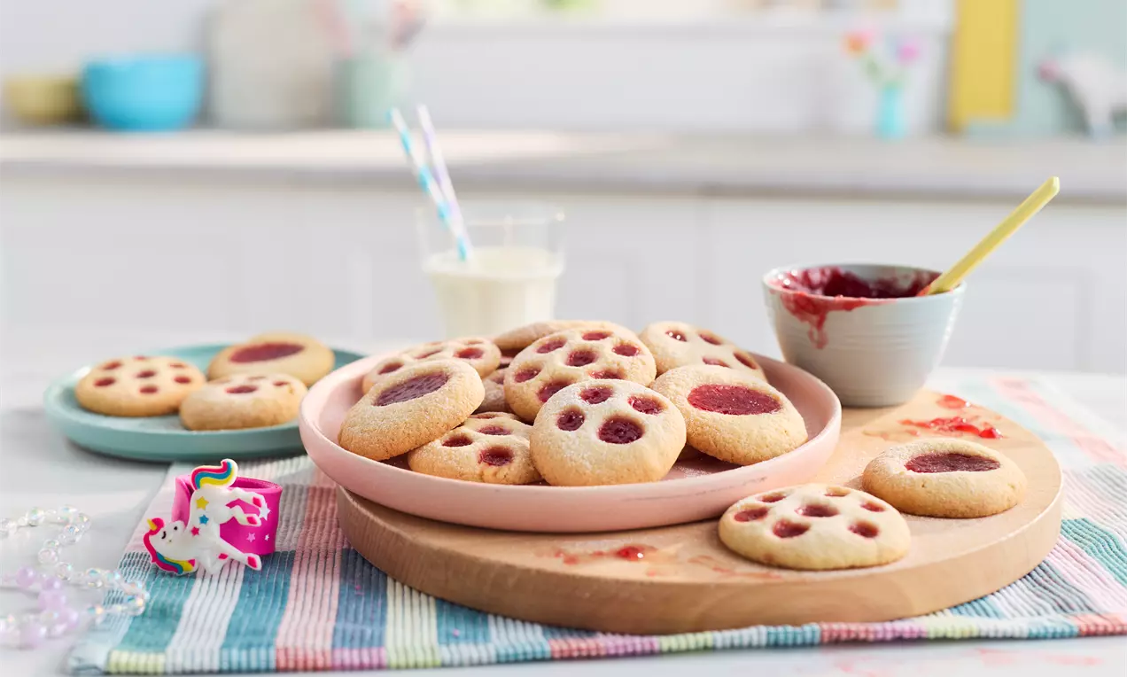 Golden, round fingerprint cookies with dimpled centers filled with red jam, arranged on pastel plates and a wooden board on a striped cloth