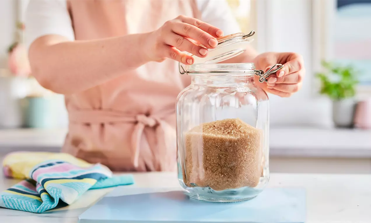 A large glass jar filled with coarse golden-brown sugar on a pastel blue surface, hands lifting the hinged lid, colourful towels nearby