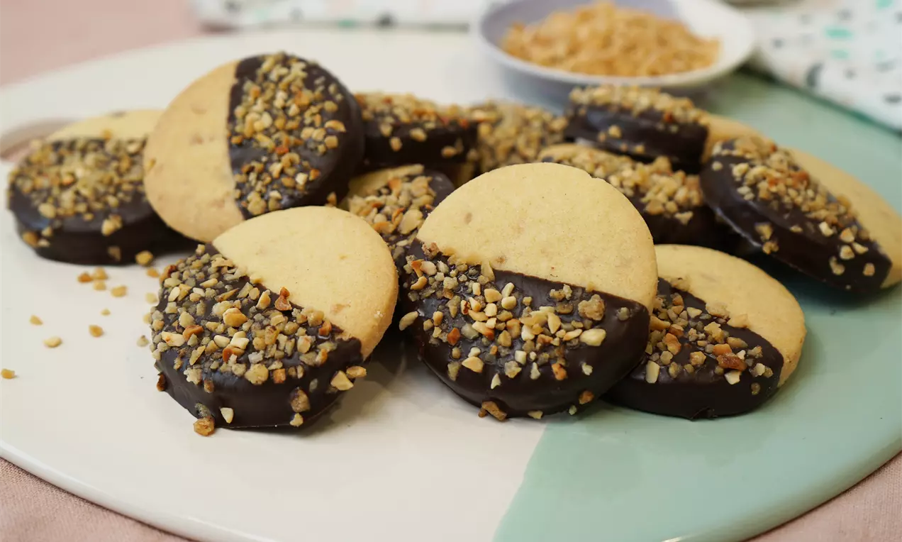 Golden round hazelnut shortbread half-dipped in glossy dark chocolate, sprinkled with chopped hazelnuts, arranged on a pastel plate