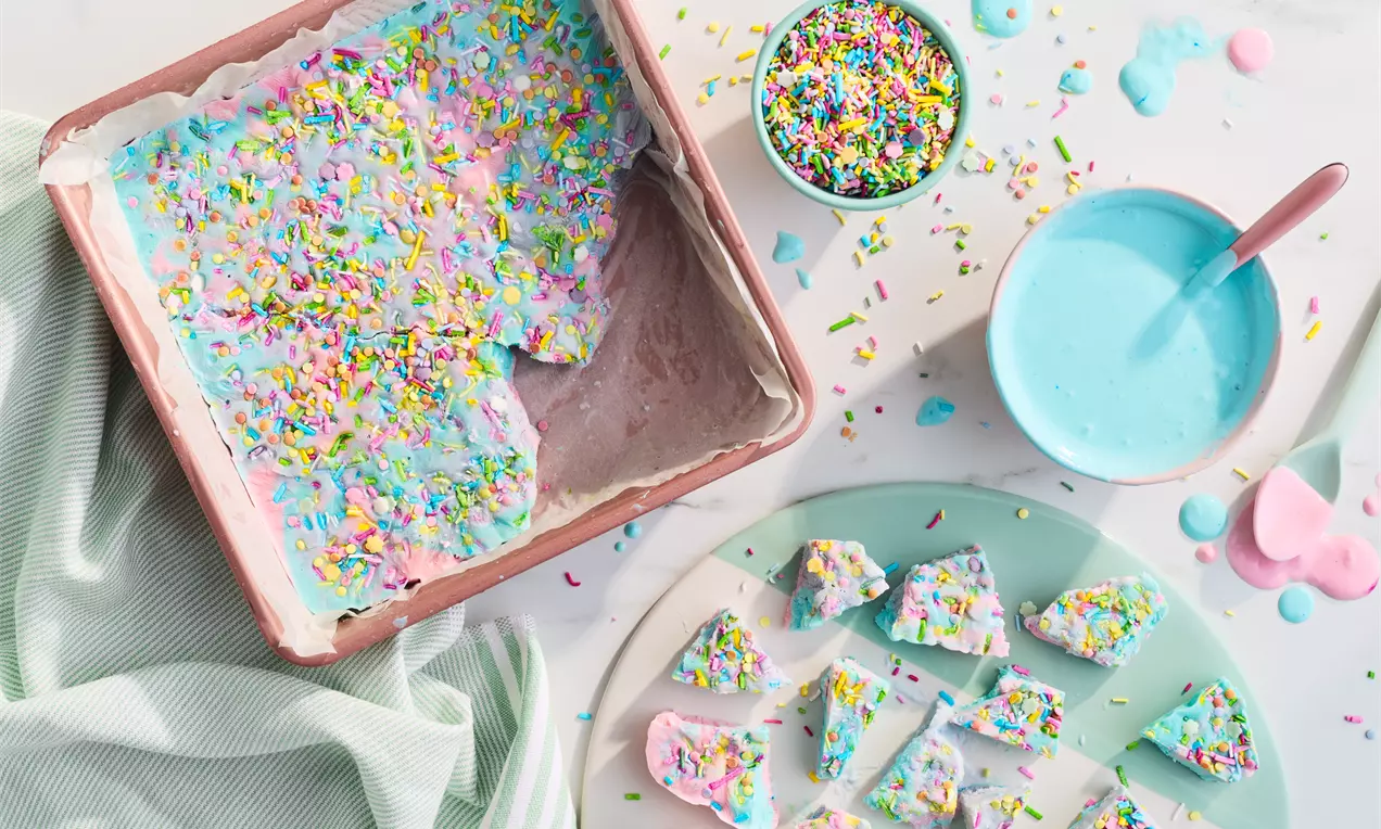 Pastel pink and white yoghurt bark topped with rainbow sprinkles, cut into irregular pieces on a tray and plate with bowls nearby