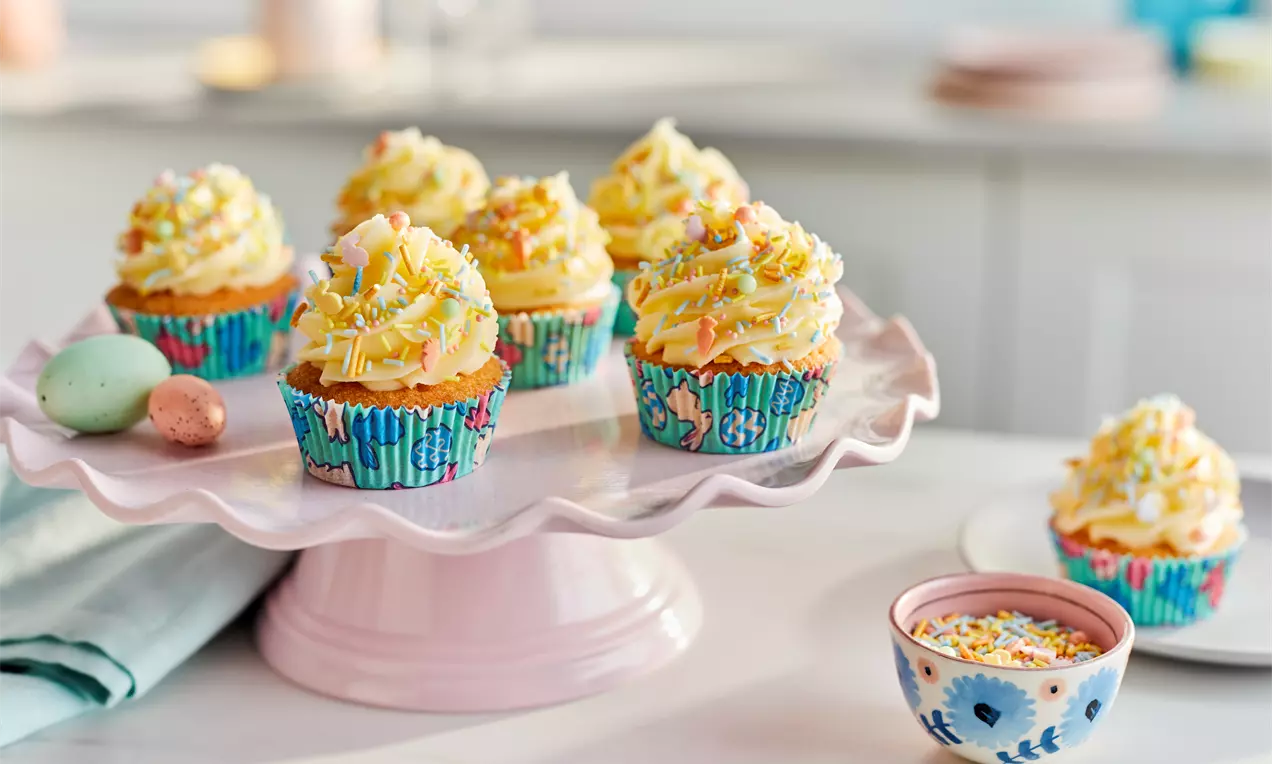Pastel yellow frosted cupcakes in vibrant floral wrappers, topped with colorful sprinkles, displayed on a pale pink scalloped stand
