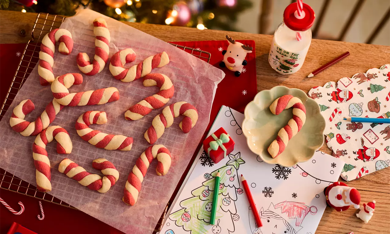 Twisted candy cane cookies in red and cream tones on a wire rack and plate, set among holiday drawings, milk, and festive decor