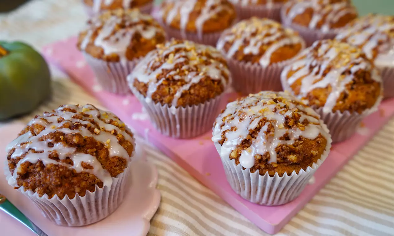 Golden-brown cinnamon roll muffins in white liners drizzled with white glaze, arranged on a pink tray atop a striped cloth, AI-labeled image