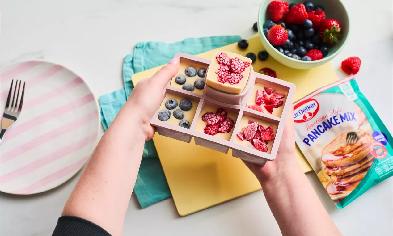 Frozen pancake block divided into compartments, each filled with berries, on a pastel background with a plate and fruit bowl nearby