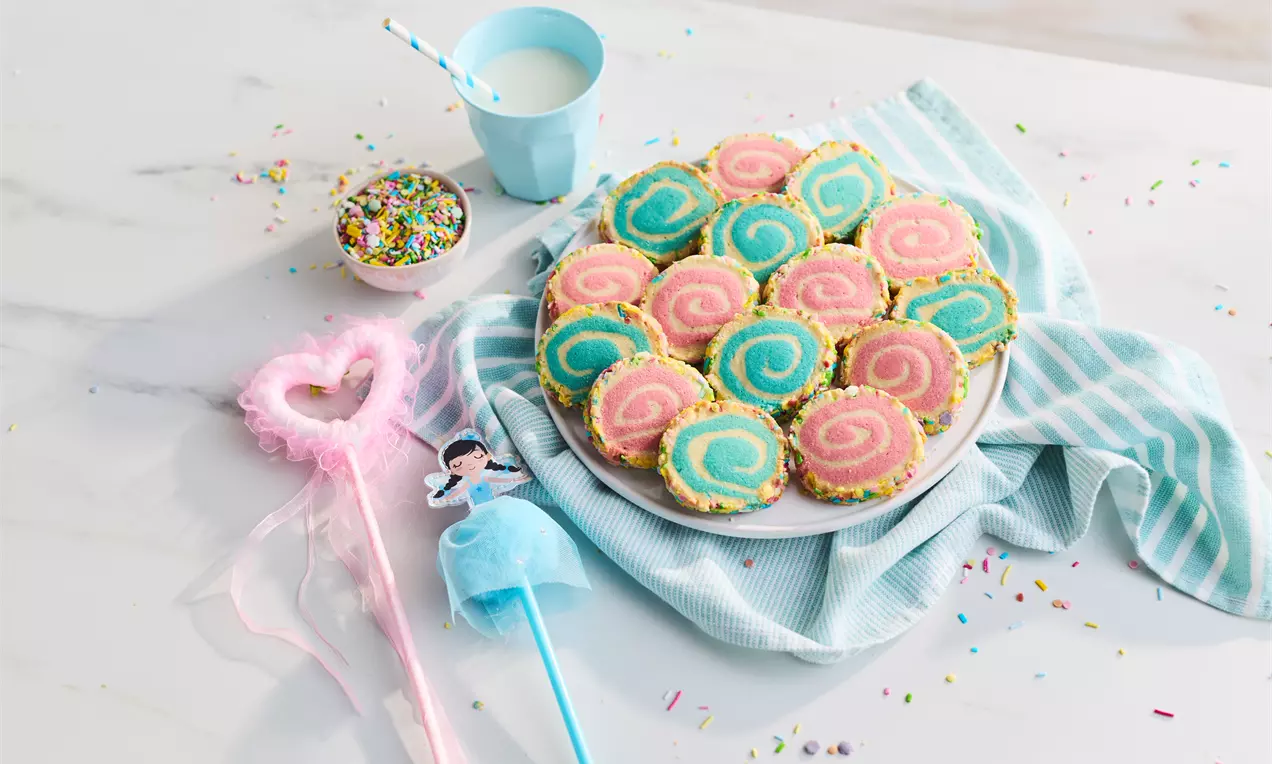 Colourful pinwheel cookies with pink and blue swirls edged in rainbow sprinkles arranged on a white plate on a striped cloth