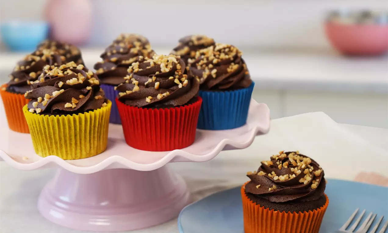 Rich chocolate cupcakes with swirls of smooth Nutella frosting, sprinkled with chopped hazelnuts, in colourful wrappers on a pink stand