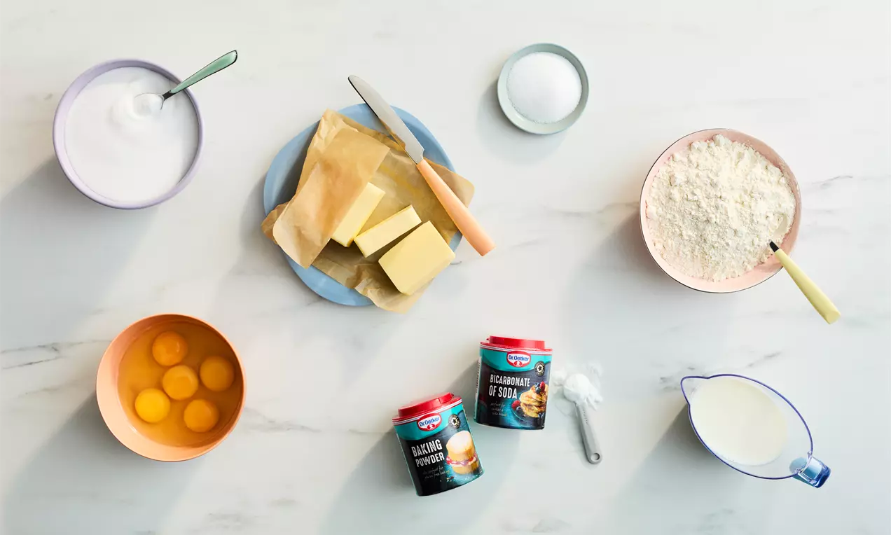 Seven baking staples displayed on a white marble surface include eggs, butter, sugar, flour, milk, and leavening agents in colorful bowls