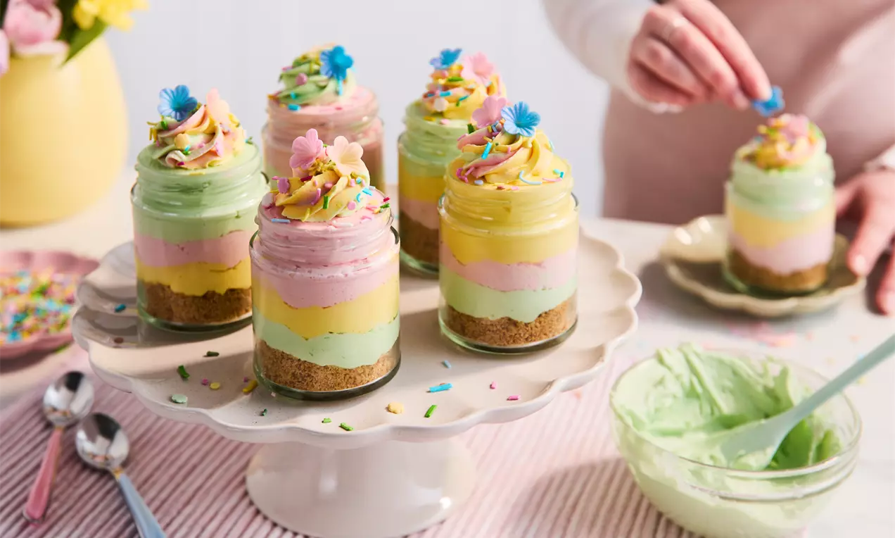 Glass jars layered with crumbly biscuit base, pastel green, pink and yellow cheesecake layers topped with swirled cream and sprinkles