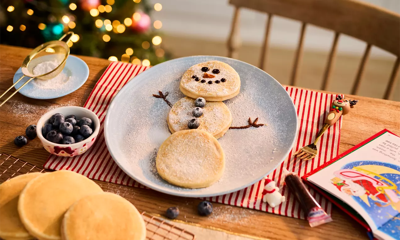 Three round golden pancakes arranged as a snowman on a light blue plate, dusted with icing sugar, with chocolate features and twig-like arms