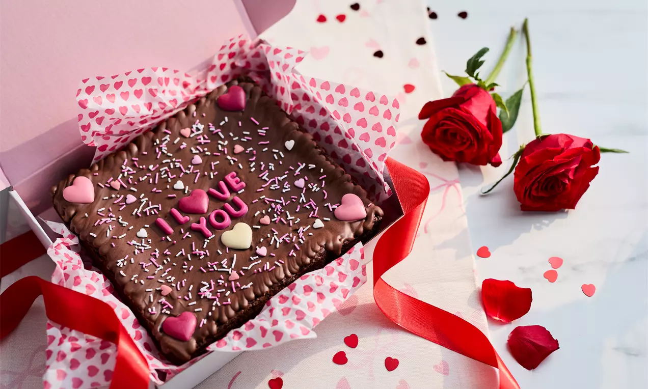 Chocolate brownies with a glossy surface, heart-shaped decorations, sprinkles, and "I love you" text inside pink heart-patterned wrapping