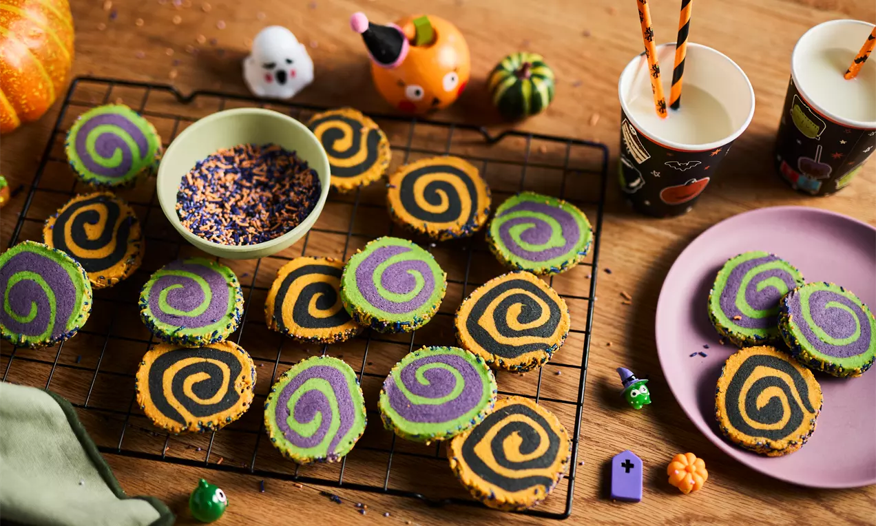 Bright Halloween pinwheel cookies with vivid black, purple, green, and orange swirls on a wire rack beside a bowl of sprinkles and festive decorations