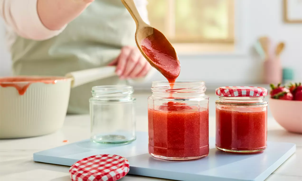 Wooden spoon pouring vibrant red jam with a smooth texture into glass jars, one with a red gingham lid, set on a light blue surface