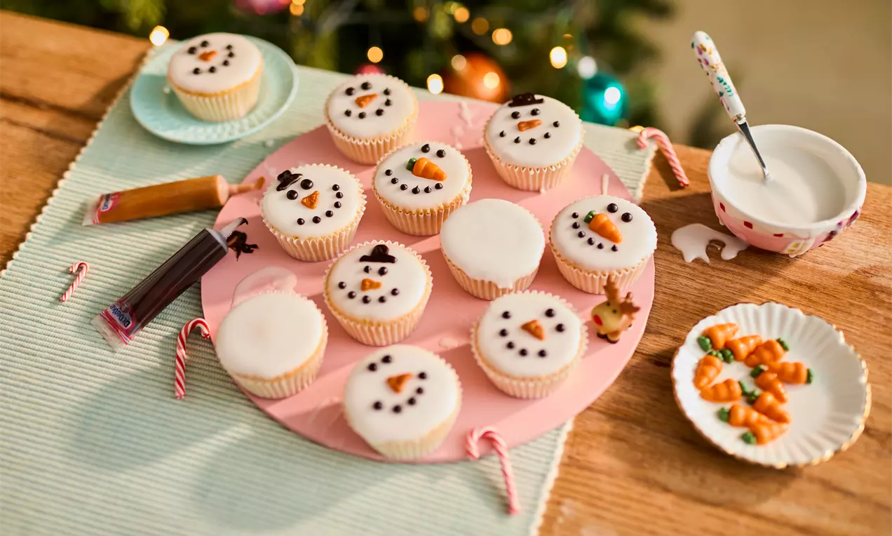 Golden cupcakes topped with smooth white frosting, decorated with snowman faces made of black and orange icing, arranged on a pink plate