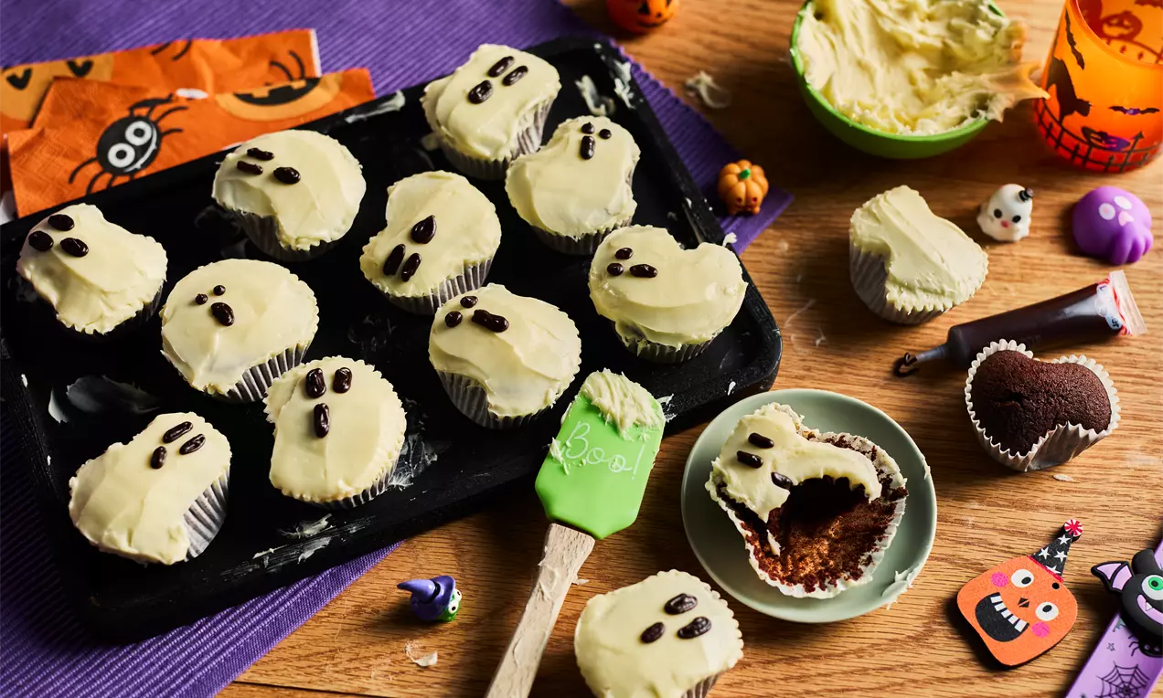 Ghost-shaped Halloween cupcakes with creamy white frosting, dark chocolate chip eyes, arranged on a black tray with festive decorations
