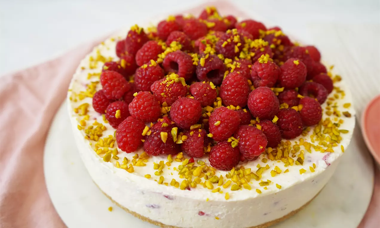 A round cheesecake with a creamy pale base and a topping of vibrant red raspberries and chopped golden-green pistachios on a marble plate