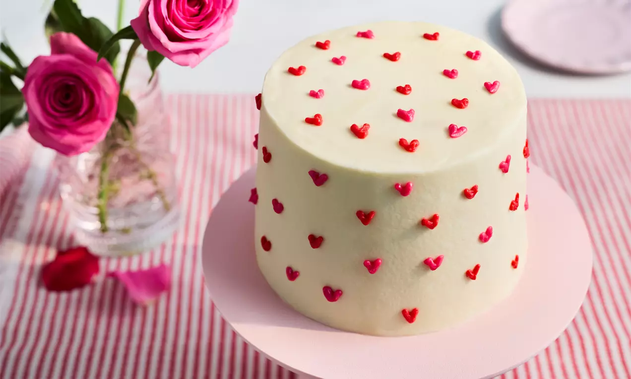 Tall round cake covered in smooth white icing, adorned with small red and pink heart decorations, on a pale pink cake stand