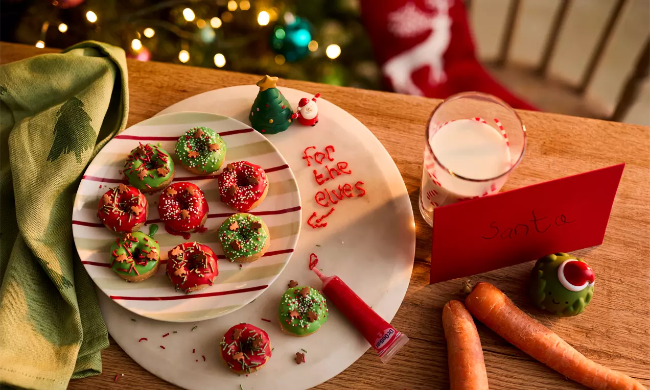 Mini doughnuts in festive red and green icing with sprinkles, arranged on a white plate with red stripes next to carrots and a note