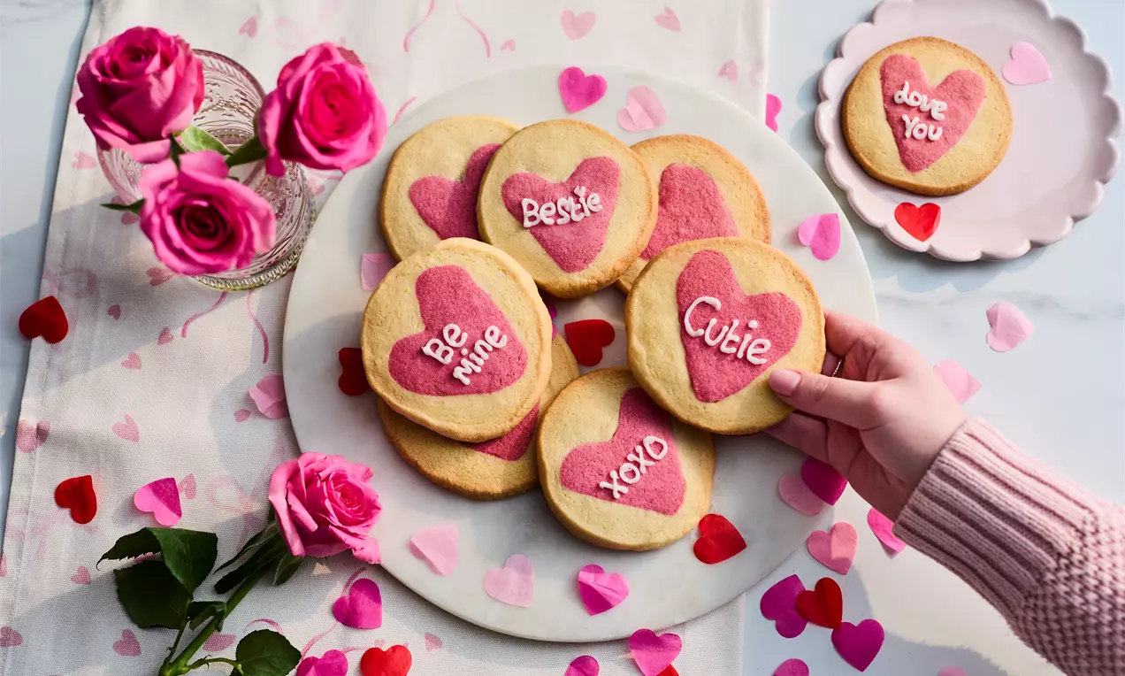 Heart-shaped cookies with pink and red centers, stamped with text, arranged on a plate surrounded by roses and scattered heart decorations