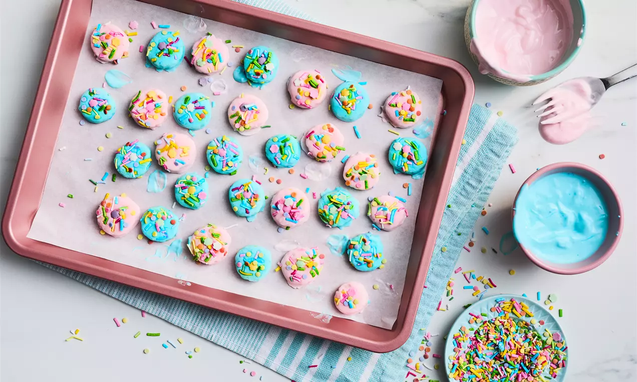Colourful banana yoghurt bites coated in pastel blue and pink yoghurt, topped with rainbow sprinkles on parchment paper in a pink tray