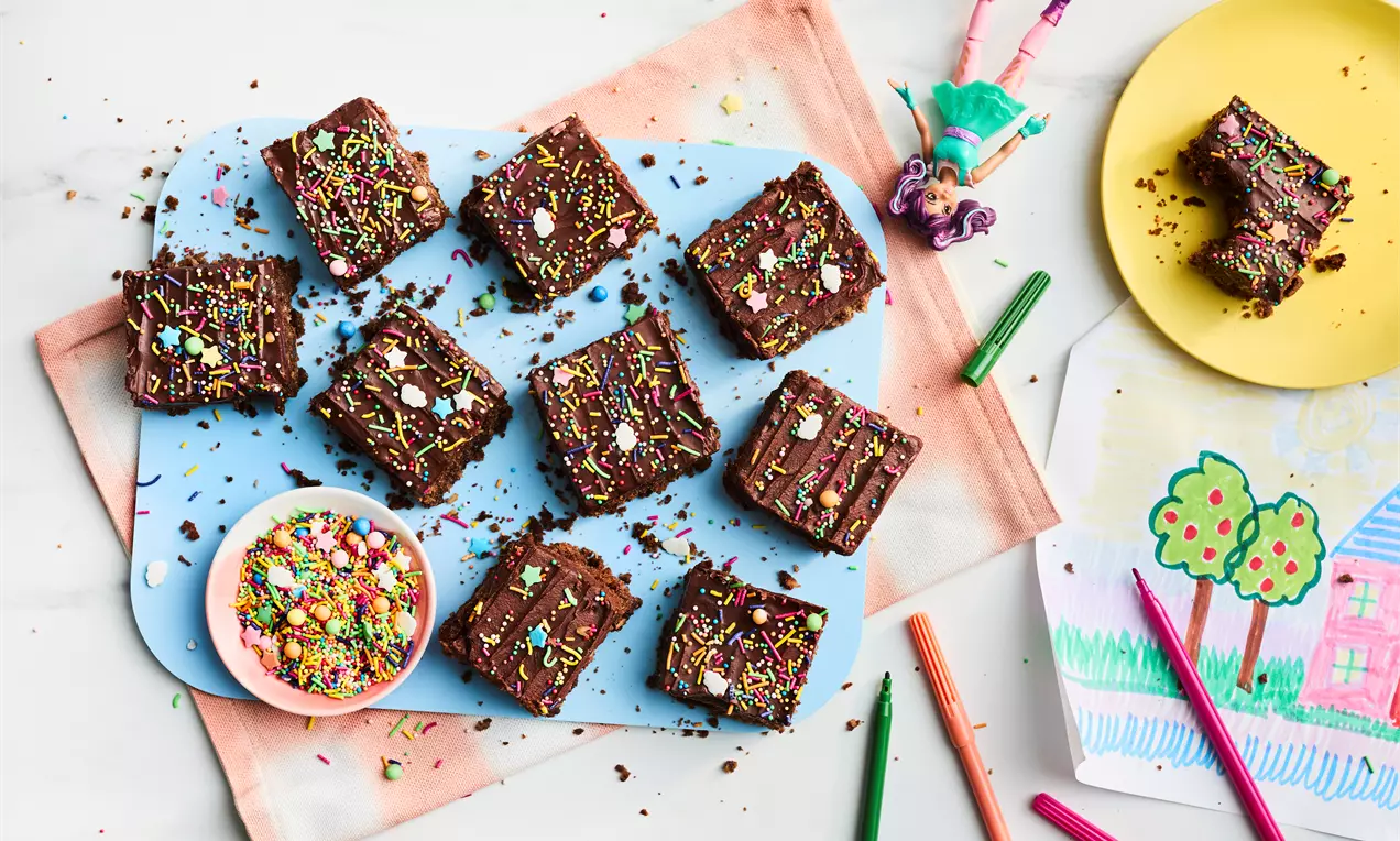 Square chocolate slices with a glossy surface, topped with colourful sprinkles, arranged on a blue board beside a bowl of sprinkles