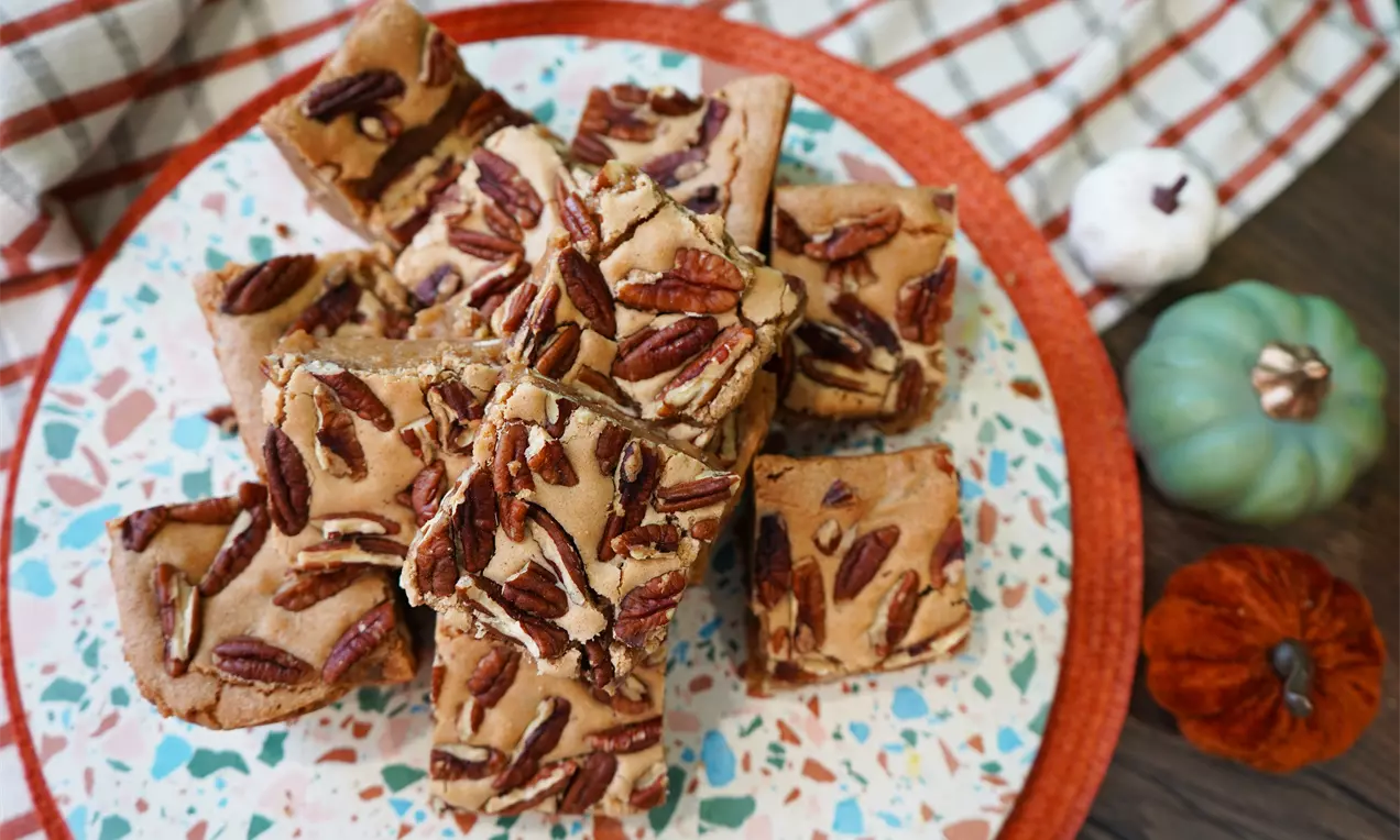 Golden maple and pecan blondie squares topped with scattered pecan halves on a colorful speckled plate with autumnal decor nearby, AI-labeled image