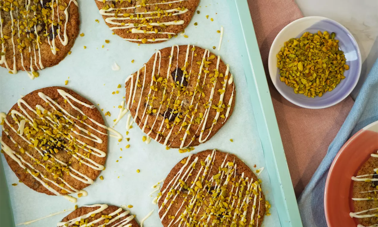 Golden brown pistachio chocolate cookies with a round shape, drizzled with white glaze and sprinkled with chopped pistachios on a blue tray