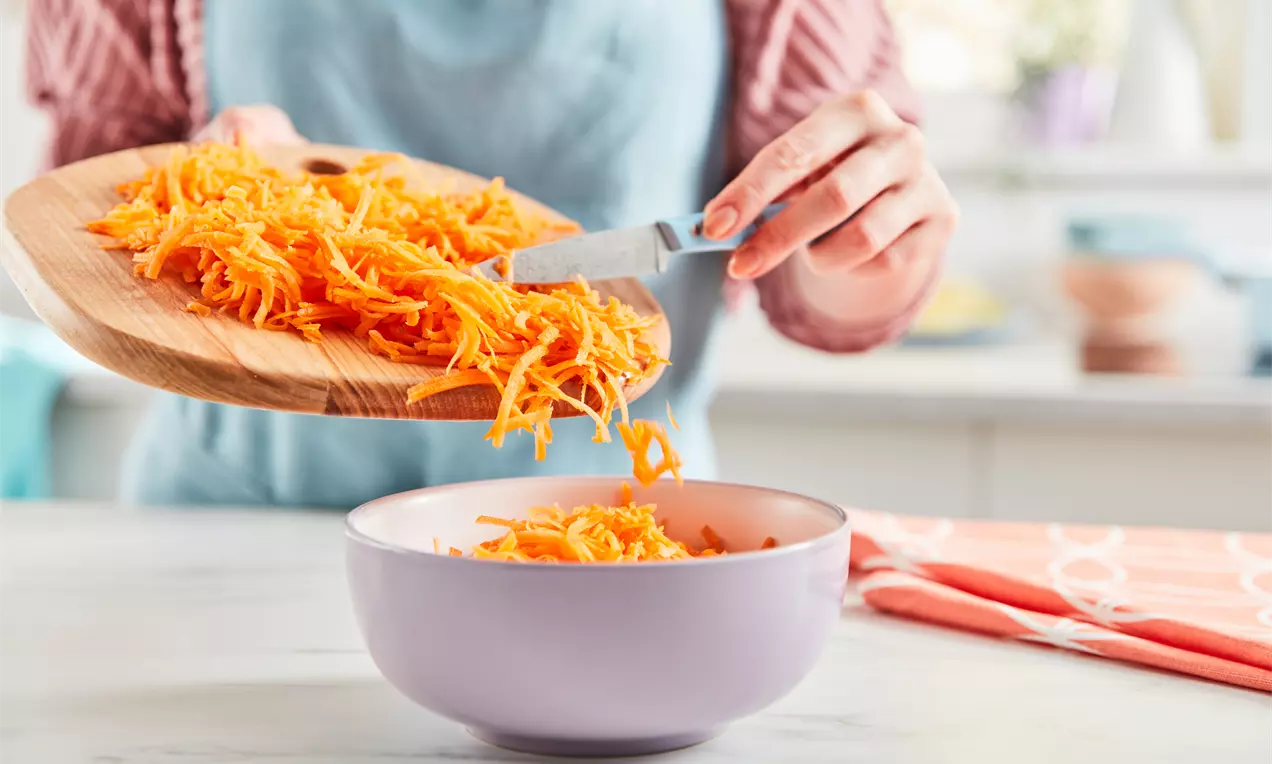 Bright orange carrot shreds with a crisp texture being transferred from a wooden board into a soft lavender bowl on a light countertop