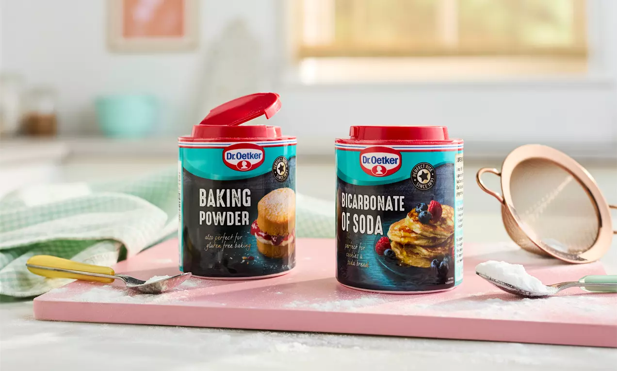 Two cylindrical tins with red lids, vibrant labels showing baked goods, placed on a pink board with loose powder and utensils nearby