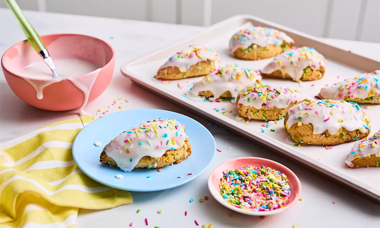 Golden triangular scones topped with glossy white icing and colourful sprinkles, arranged on a tray and a blue plate with sprinkle dishes nearby