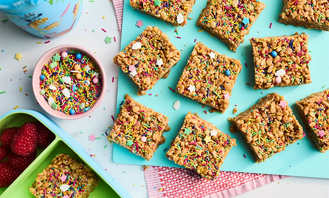 Golden-brown Rice Krispie treats cut into squares, topped with colorful sprinkles and candies, arranged on a light blue tray