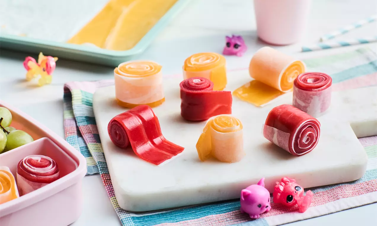Colourful fruit roll-ups in red, orange, and yellow, neatly rolled and arranged on a white marble slab with small toy decorations